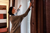 istock Young beautiful Muslim woman is opening blinds on a window in a living room. 1361274133