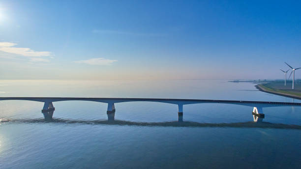 The Zeelandbrug during daytime. The sky is clear blue and the water is peaceful, because of this the bridge and the sky are reflected in the water. On the coastline are two big wind turbines.