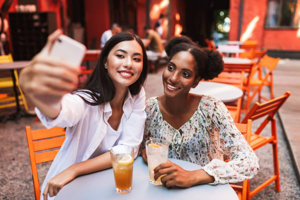 istock Two young ladies sitting at table with cocktails taking photos on cellphone happily while spending time together in cozy courtyard of cafe 1361286889
