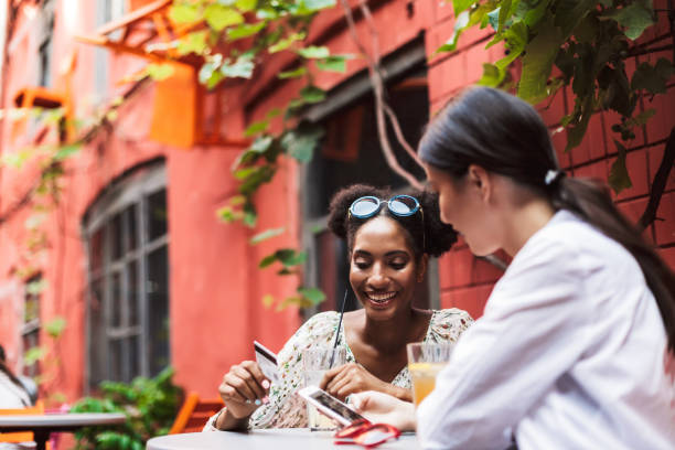 istock Pretty smiling girls using cellphone and credit card while happily spending time together in cozy courtyard of cafe 1361287922