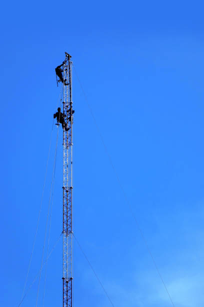 A spectacular silhouette of a man working on a metal tower against a cloudy sky. Loosely hanging cables indicate construction, repair or telecommunication work
