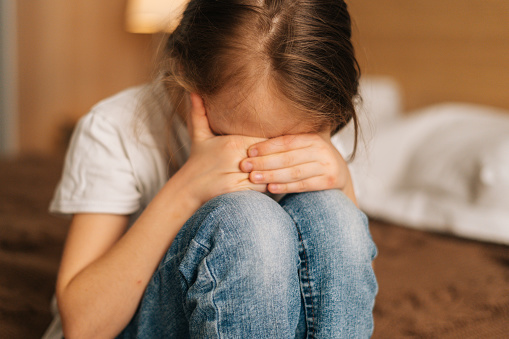 Close-up of disappointed little girl hugging knee, sobbing with head bowed and crying sitting on bed in bedroom. Stressed child feeling pressure, thinking of kid problems. Concept of child depression.