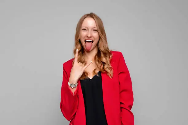 Portrait of nice, attractive, crazy young blonde woman, showing tongue out and rock-n-roll sign, standing in red blazer against gray background