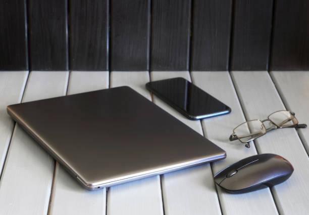 A thin silvery laptop, a computer mouse, a smartphone and glasses lie on a wooden surface. Scandinavian ecological workplace concept. Coworking space and distant teleworking