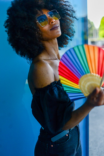 portrait of a beautiful african american girl wearing sunglasses and usign a rainbow colored fan in summer,  lgbtq+ pride concept