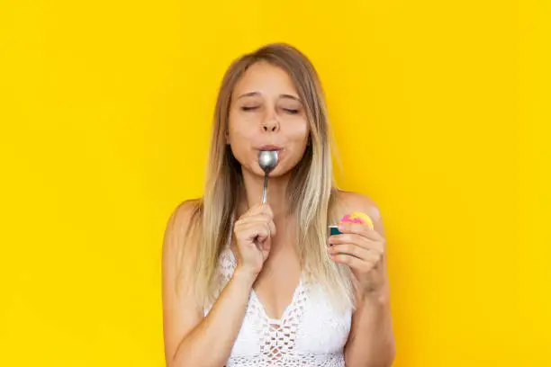 A young beautiful caucasian blonde woman with her eyes closed enjoys the taste of sorbet, licking a spoon and holding a cream bowl with ice cream isolated on a color yellow background. Summer concept