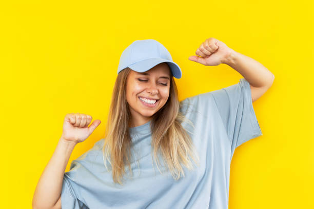 青い帽子をかぶった若い笑顔のブロンドの女性は、ニュースや宝くじが色の黄色の背景に勝つことに満足しています - oversized ストックフォトと画像