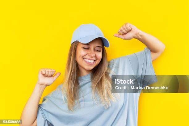 A Young Smiling Blonde Woman In A Blue Cap Is Happy About The News Or Lottery Win On Color Yellow Background Stock Photo - Download Image Now
