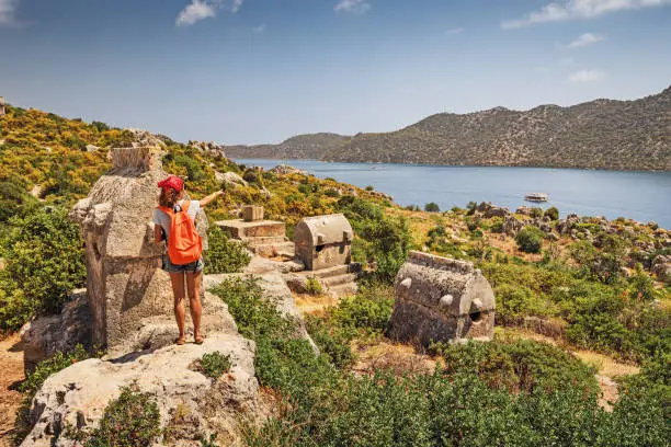 Photo of Girl traveler exploring ancient ruins of Lycian necropolis city with fascinating Tombs and sarcophagus near Kekova island in Turkey. Historical sights and archeology concept