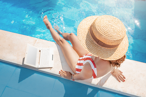 Woman in a swimsuit is sitting by the pool and surfing the internet social networks by her laptop pc with blank screen. Studying at online university and remote work