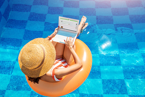 Woman in a swimsuit floating in inflatable circle in swimming pool and surfing the internet websites at her laptop pc. Leisure weekend and remote freelance work concept
