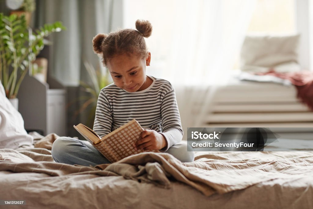 Little African American Girl Reading Book on Bed Full length portrait of cute African-American girl reading book while sitting on bed in cozy interior, copy space Child Stock Photo
