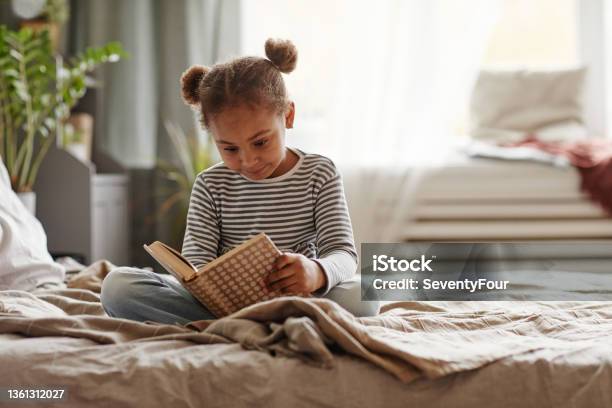 Little African American Girl Reading Book On Bed Stock Photo - Download Image Now - Child, Reading, Book