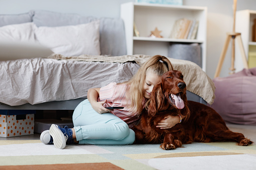 Full length portrait of cute blonde girl playing with dog on floor in cozy home interior , copy space