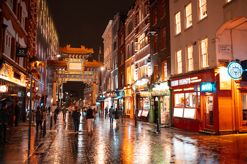 Wet pedestrian street with people walking towards Chinese gate, illuminated Chinese restaurants on both sides, Gerrard Street in Soho - London at night