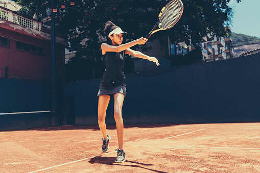 A young black woman playing tennis on a clay sports court. A woman in black sportswear and a white visor holding a tennis racket. A tennis player training by attacking serves at a private club