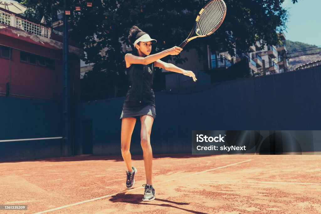 A tennis player making a serve A young black woman playing tennis on a clay sports court. A woman in black sportswear and a white visor holding a tennis racket. A tennis player training by attacking serves at a private club Tennis Stock Photo