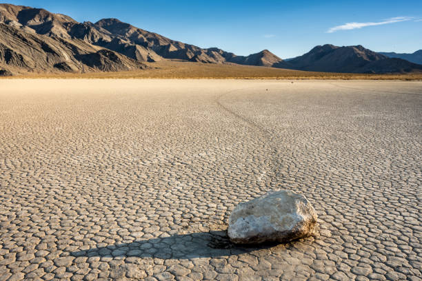 The terrain is a mix of golden sand dunes, rugged rocky formations, and salt flats glistening under the intense sun. The sky is a brilliant blue, contrasting with the warm earthy tones of the valley below. In the distance, jagged mountains rise, their peaks softened by a hazy mirage. The extreme conditions of this iconic location make it one of the most unique and mesmerizing places in the world