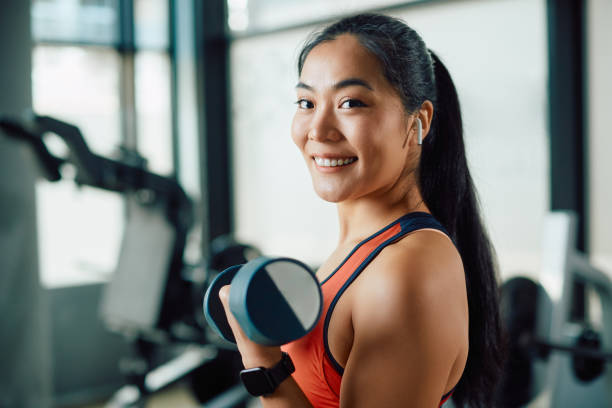 feliz mujer atlética asiática haciendo ejercicio con pesas de mano en un gimnasio y mirando a la cámara. - culturismo fotografías e imágenes de stock