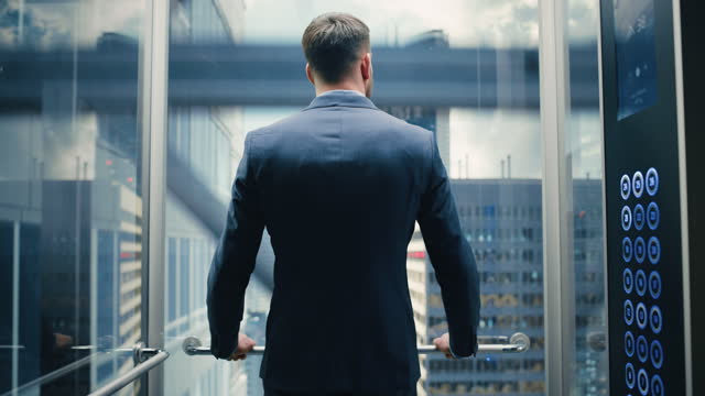 Successful Businessman in a Suit Riding Glass Elevator to Office in Modern Business Center. Young Male Looking at Modern Downtown Skyscrapers Out of the Panorama Window in the Lift.