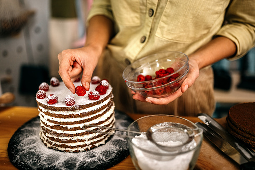 Selective focus of hands of young women adding juicy raspberries on cake with powdered sugar and icing for garnish at kitchen counter