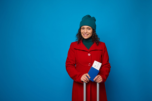 Young smiling woman wearing bright red coat and green warm woolen hat, poses with suitcase, passport and boarding pass in her hands. Travel, air flight journey concept on blue background, copy space