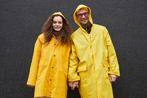 Horizontal medium shot of Caucasian man and woman wearing yellow raincoats holding hands looking at camera