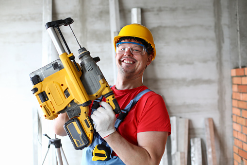 Portrait of cheerful happy carpenter holding big electrical machine. Smiling worker showing practical device for work. Foreman in uniform. Construction site and renovation concept