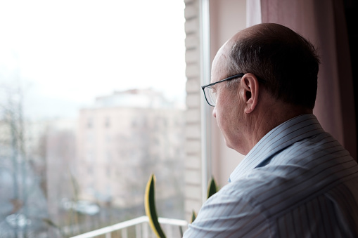 Elderly man looking out window with lonely expression