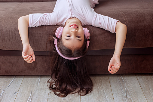 A funny playful cute girl in cozy pink home clothes and pink headphones lies hanging from a brown sofa, smiling