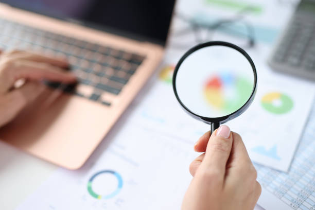 istock Woman holding magnifying glass in hands over documents with graphs and charts and working on laptop 1361402273