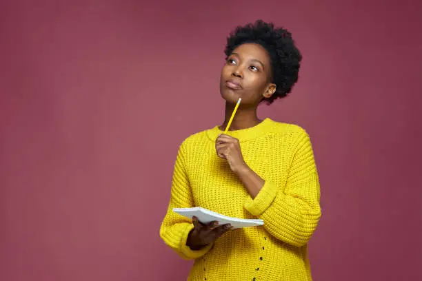 Photo of Stylish pensive african american girl pondering idea to write into notebook, thinking over business plan, to-do list