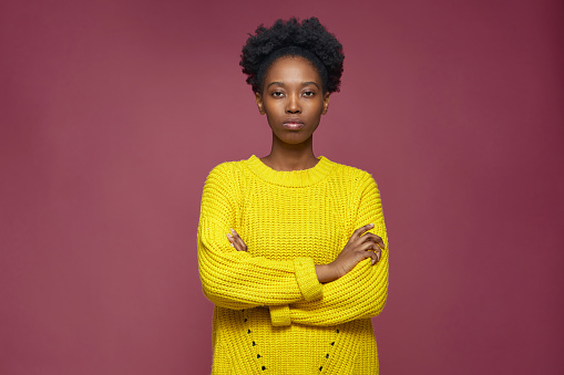 Serious stylish confident young african american girl standing with folded hands, looking at camera on pink background. High quality photo