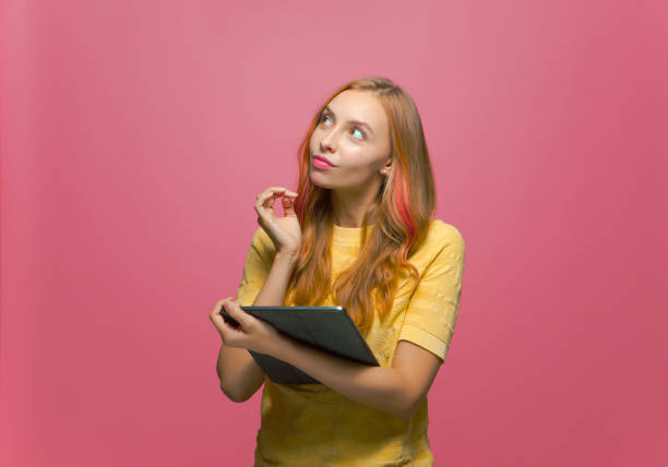 Modern dreaming young girl with tablet gadget looking up, thinking, create ideas, content for social network on pink background. High quality photo