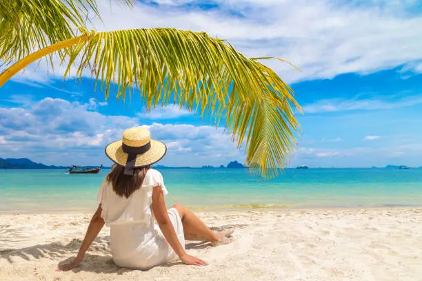 Young beautiful woman wearing white dress and straw hat is sitting under palm leaf on a tropical beach