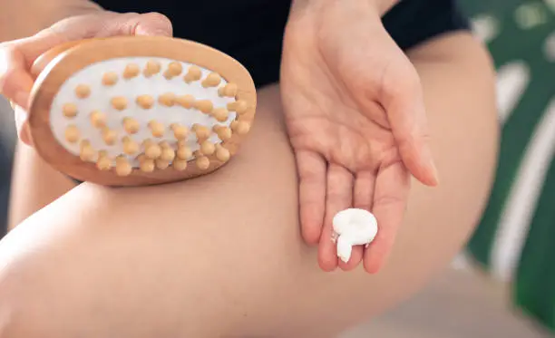 Close-up of anti-cellulite massage brush and body cream in female hands on a blurred background.