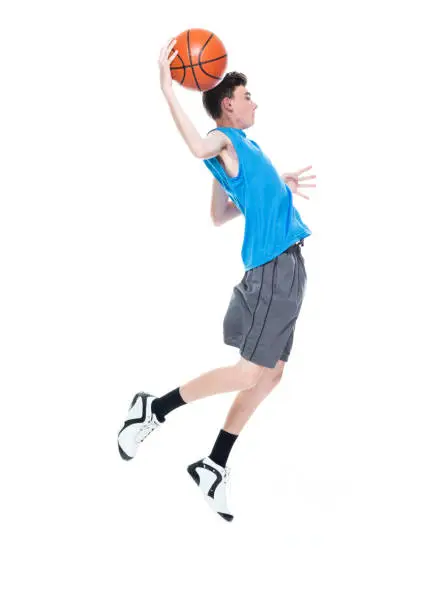 Side view of aged 12-13 years old caucasian male basketball player mid-air in front of white background who is excited and holding basketball - ball and using sports ball