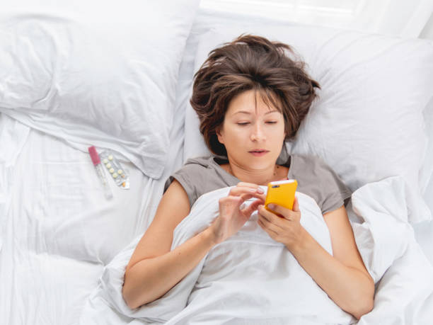 Top view on sick woman lying in bed with smartphone. Thermometer and pills on white bed linen. Internet surfing. stock photo