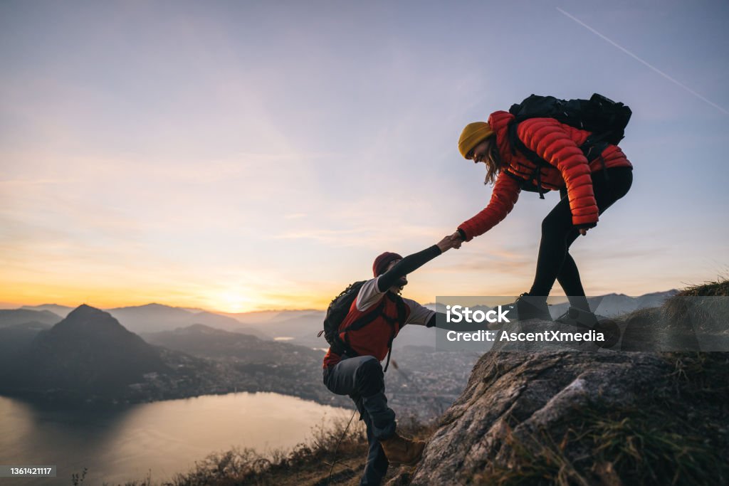 Hiking couple climb up mountain ridge Young woman offers man a hand up, in the Swiss Alps Teamwork Stock Photo