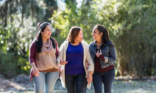 A senior Hispanic woman walking in the park, conversing with her two adult daughters on a sunny autumn day. The mother, in her 60s, is walking in the middle between her children, who are in their 40s.