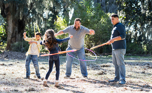 Two Hispanic brothers in their 30s playing at the park with their children, a 7 year old boy and 9 year old girl. They are standing in a circle with plastic hoops. The view is from over the girl's shoulder as she shows her father, cousin and uncle how to swing the hoop around her waist.