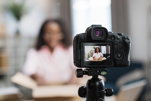 African american woman unboxing parcel with ordered goods while sitting at home and recording video blog. Female influencer using modern digital camera for creating new content.
