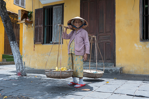 Hoi An, Vietnam - July 01, 2020 : Vietnamese woman in a straw hat with a basket of fruits on a street market in the old city against the background of the old yellow wall in Hoi An, Vietnam