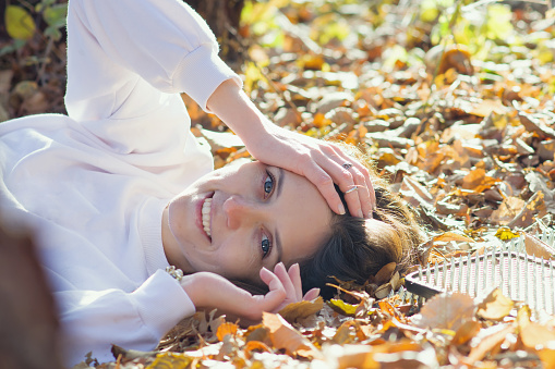 Happy young woman lies next to sadhu yoga board on the ground with autumn foliage in the park. A girl in white clothes smiles happily, looks at the camera, lying on the ground in the forest.