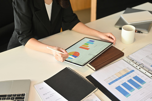 Top view image of businesswoman hand using a stylus pen and digital tablet with graphic charts on screen at the cluttered office desk.