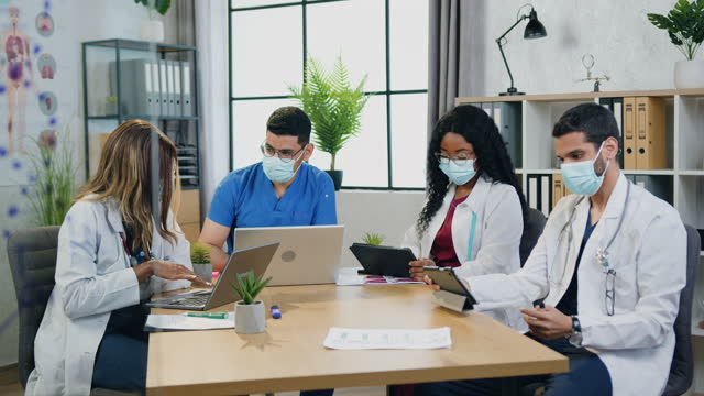 Experienced confident serious mixed race medical team in protective masks brainstorming together in consultation room in hospital,work during covid-19 pandemia