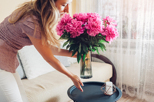 Woman puts vase with peonies flowers on table. Housewife taking care of coziness at home. Interior and spring decor