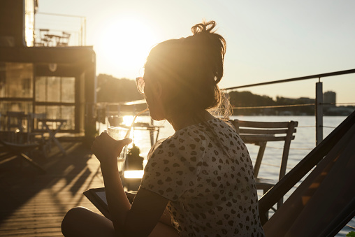 Young woman sitting in cafe and watching the river at summer sunset. Retro style image.