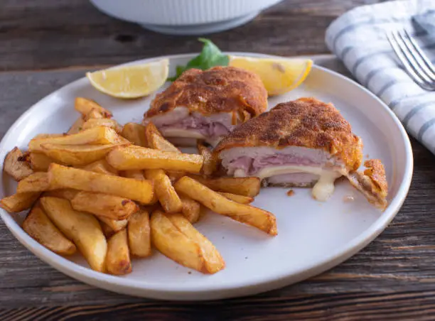 Fresh and homemade cooked cordon bleu made with chicken breast and filled with ham and cheese. Served with french fries and lemon on a plate.