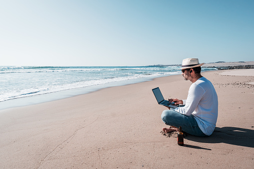 man sitting on the beach with a laptop alone doing telecommuting or remote work and drinking beer